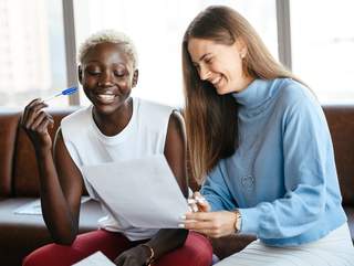 Two women sit in front of a window and look together at a paper. One woman points to a place in the text with a pen, the other woman looks at it. She also has a pen in her hand. Both women laugh.